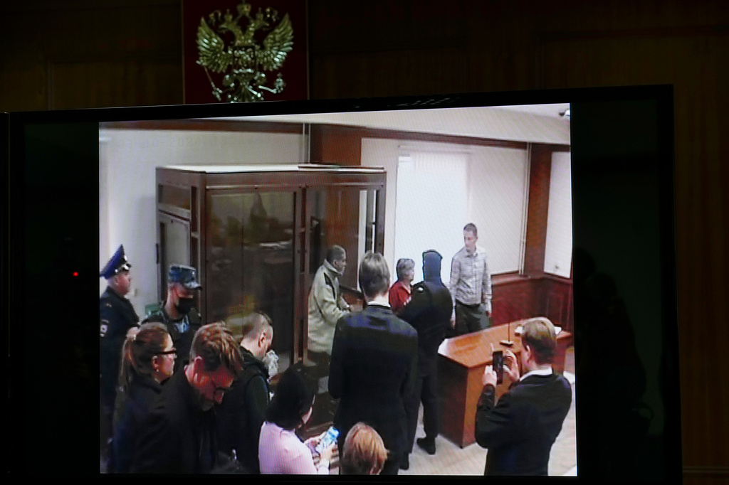 Photo of a screen showing stephen hubbard standing in a glass box in a courtroom in russia surrounded by people